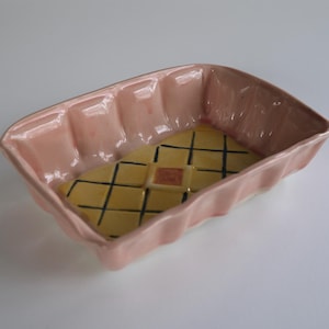 May include: A rectangular, pink ceramic dish with a yellow interior featuring a black grid pattern and a central square. The dish has a wavy, ridged edge and is set against a white background.