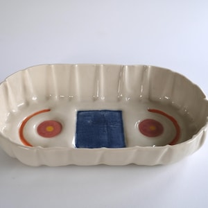 Puede incluir: Fuente de cerámica color crema con borde acanalado. El interior presenta un cuadrado azul y dos diseños circulares rojos y amarillos. Una pieza única, hecha a mano.