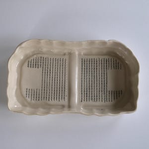 Pode incluir: Uma travessa de servir de cerâmica creme com borda ondulada. O interior apresenta duas secções retangulares com um padrão de linhas pretas verticais. A travessa está sobre uma superfície branca.