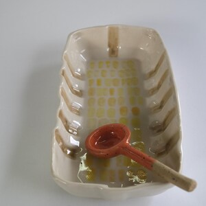 Puede incluir: Una fuente de cerámica con una pequeña cuchara naranja. La fuente es rectangular con bordes redondeados y un esmalte beige. Tiene rayas marrones y un patrón de cuadros amarillos y blancos. La cuchara es naranja con un mango beige.