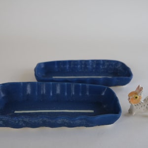 Puede incluir: Dos platos rectangulares de cerámica azul con líneas blancas en la parte inferior. Los platos tienen una superficie texturizada y son ligeramente ondulados. Una pequeña figurita de cerámica blanca de un conejo con ojos naranjas está en primer plano.