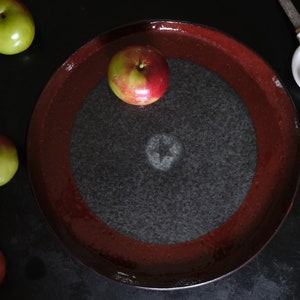 Op de afbeelding: Een rood en zwart keramisch bord met een enkele rode appel erop. Er zijn verschillende groene en rode appels rond het bord.