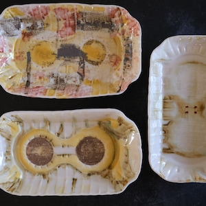 Peut inclure: Trois plats rectangulaires en céramique avec des motifs différents. Le premier plat a un motif abstrait jaune, rouge et brun. Le deuxième plat a un motif jaune et brun avec deux indentations circulaires. Le troisième plat est blanc avec un motif brun.