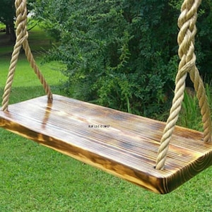 May include: A wooden swing with a natural wood finish and rope hanging from the top. The swing is hanging in a grassy area with green trees in the background.