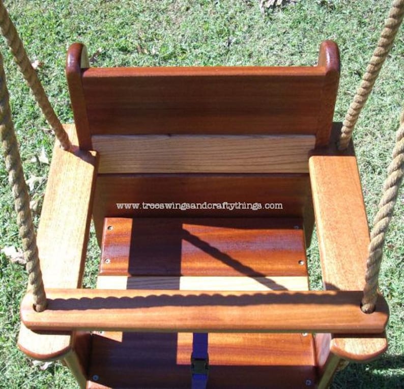 May include: A wooden baby swing with a natural finish, featuring a seat, backrest, and armrests. The swing is suspended by thick, beige ropes. The text "www.treeswingsandcraftythings.com" is visible on the swing.