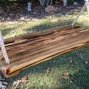 May include: A wooden swing with a rich brown finish and visible wood grain. The swing is rectangular with rounded corners and is suspended by thick, off-white ropes. The swing is designed for outdoor use and is set against a backdrop of grass and foliage.