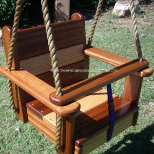 May include: A wooden baby swing with a natural finish, featuring a seat, back, and armrests. The swing is suspended by thick, woven ropes. A blue strap with a black buckle is visible. The website address www.treeswingsandcraftthings.com is printed on the swing.