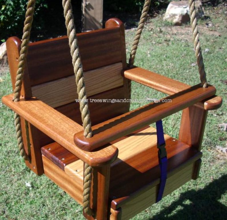 May include: A wooden baby swing with a natural finish, featuring a seat, back, and armrests. The swing is suspended by thick, woven ropes. A blue strap with a black buckle is visible. The website address www.treeswingsandcraftthings.com is printed on the swing.