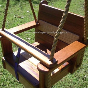 May include: A wooden baby swing with a high back and armrests. The swing is made of stained wood and features a seat with a purple safety strap. Thick rope is used for the swing's suspension. The swing is designed for outdoor use.