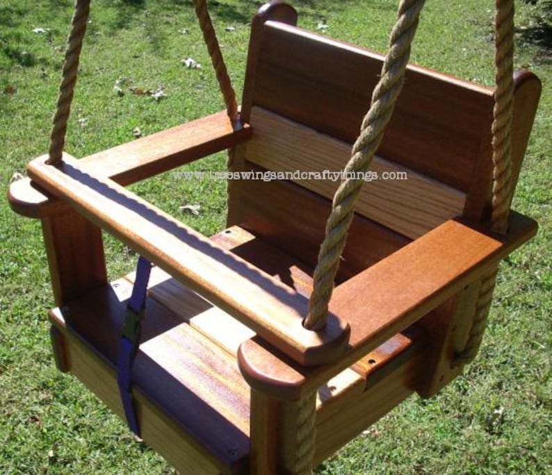 May include: A wooden baby swing with a high back and armrests. The swing is made of stained wood and features a seat with a purple safety strap. Thick rope is used for the swing's suspension. The swing is designed for outdoor use.