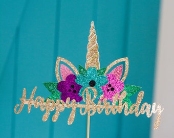 Puede incluir: Topper de pastel de purpurina dorada con una cabeza de unicornio y flores, y las palabras "Feliz cumpleaños".