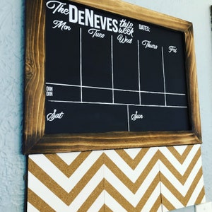 May include: A black chalkboard with a wooden frame, divided into seven sections for each day of the week. The chalkboard has the text "The Deneves this week" written in white cursive. The days of the week are labelled "Mon", "Tues", "Wed", "Thurs", "Fri", "Sat", and "Sun". The chalkboard is mounted on a wall with a corkboard underneath.