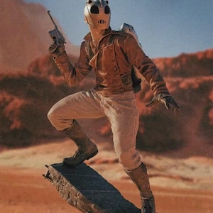 Peut inclure: Un casque argenté et noir avec une visière et une grande ouverture d'œil ronde noire. Le casque est porté par une personne vêtue d'une veste en cuir marron et d'un pantalon blanc. La personne se tient debout sur un morceau de béton cassé, tenant un pistolet dans sa main droite. L'arrière-plan est un paysage désertique.