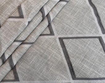 May include: Close-up of a folded fabric with a geometric pattern. The fabric is a light gray color with darker gray geometric shapes. The shapes are outlined with a darker gray border. The fabric has a textured appearance.