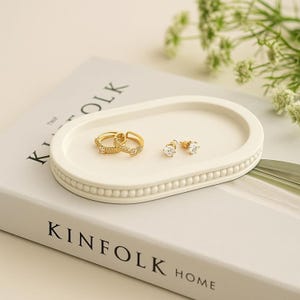 May include: A white oval jewelry tray with a beaded edge holds two gold hoop earrings and a pair of gold stud earrings with clear stones. The tray rests on a book with the text "KINFOLK HOME".