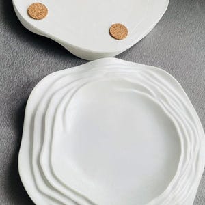 May include: Two white ceramic serving dishes. The top dish is oval with four cork feet. The bottom dish is round with a wavy, layered design. Both dishes are on a gray surface.