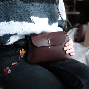 May include: A brown leather crossbody bag with a flap closure and a gold metal accent. The bag is worn over a person's shoulder and is being held by their hand.