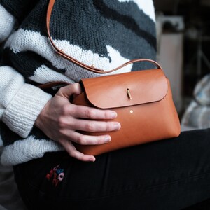 May include: A brown leather crossbody bag with a flap closure and a gold metal accent. The bag is being held by a person wearing a white and black striped sweater and black jeans.