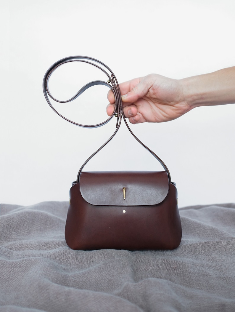 May include: A brown leather handbag with a flap closure and a gold metal clasp. The bag has a long strap that can be worn over the shoulder.