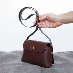 May include: A brown leather crossbody bag with a gold clasp and a thin strap. The bag is sitting on a light brown fabric surface.