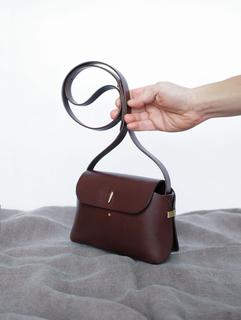 May include: A brown leather crossbody bag with a gold clasp and a thin strap. The bag is sitting on a light brown fabric surface.