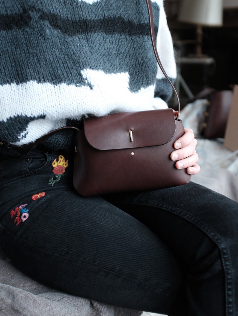 May include: A brown leather crossbody bag with a flap closure and a gold metal accent. The bag is worn over a person's shoulder and is being held by their hand.