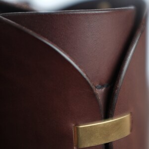 May include: Close-up of a brown leather bucket with a gold metal accent. The bucket has a unique, folded design.