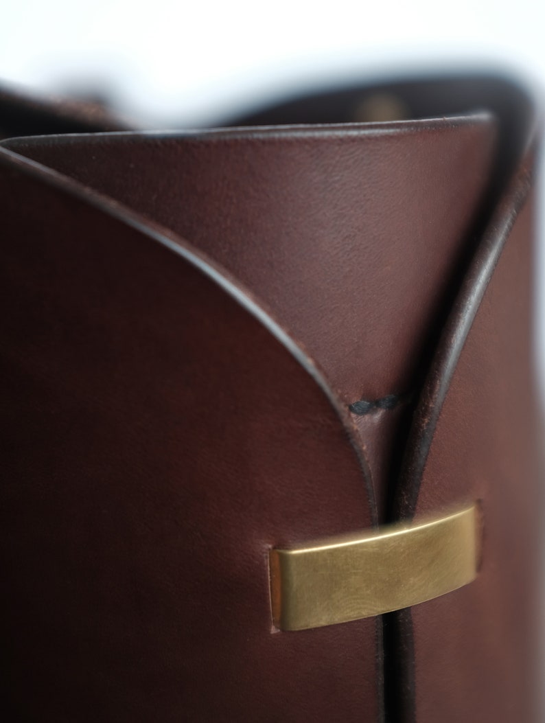 May include: Close-up of a brown leather bucket with a gold metal accent. The bucket has a unique, folded design.