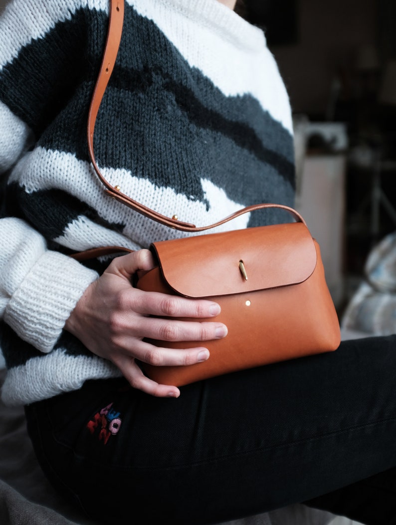 May include: A brown leather crossbody bag with a flap closure and a gold metal accent. The bag is being held by a person wearing a white and black striped sweater and black jeans.