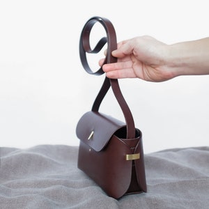 May include: A brown leather crossbody bag with a gold clasp and a long strap. The bag is sitting on a gray fabric surface.