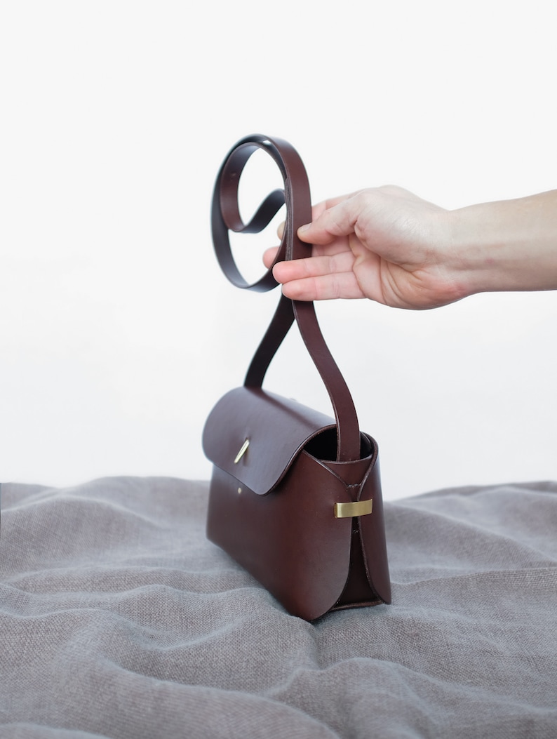 May include: A brown leather crossbody bag with a gold clasp and a long strap. The bag is sitting on a gray fabric surface.