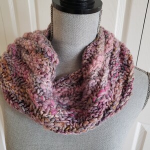 May include: A pink, purple, and brown knitted infinity scarf. The scarf is made with a chunky yarn and has a textured stitch pattern.