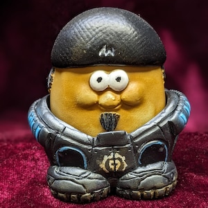 Puede incluir: Un personaje de patata marrón que lleva una boina negra y un traje espacial negro y azul con un símbolo de engranaje en el pecho.