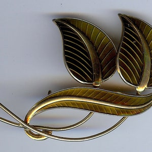 Peut inclure: Une broche dorée avec trois feuilles stylisées. Les feuilles sont émaillées dans des tons de vert et de noir.