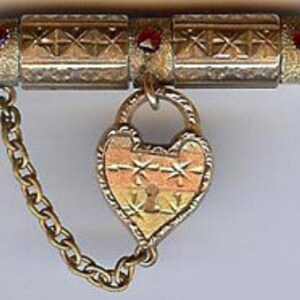 May include: A gold-toned locket with a heart-shaped design and a small keyhole. The locket is attached to a chain and has two red gemstones on the top.