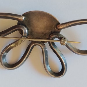 Puede incluir: Un broche de plata con un centro grande, redondo y plateado y un delicado alfiler dorado. El broche tiene forma de flor estilizada con m&uacute;ltiples bucles.