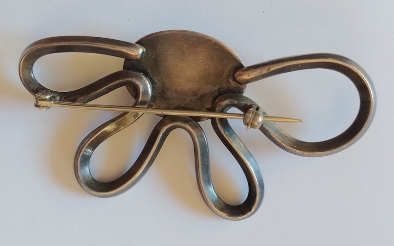 Puede incluir: Un broche de plata con un centro grande, redondo y plateado y un delicado alfiler dorado. El broche tiene forma de flor estilizada con m&uacute;ltiples bucles.