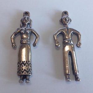 May include: Two silver-toned metal pendants. One pendant depicts a woman with a long skirt and arms outstretched. The other pendant is a man with arms outstretched and wearing pants. Both pendants have a loop at the top for hanging.