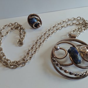 Può includere: Una collana d'argento con un ciondolo con una pietra blu e un anello d'argento con una pietra blu.