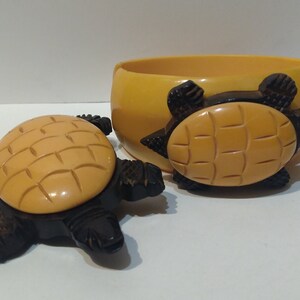 May include: A set of two vintage yellow and black Bakelite turtle-shaped jewelry pieces. The larger piece is a bangle bracelet with a turtle design on the front. The smaller piece is a brooch with a turtle design.