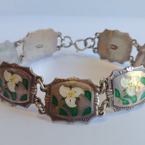 May include: A silver bracelet with white and green enamel flowers. The bracelet is made of linked sections, each with a different design. The back of the bracelet has a maker's mark.