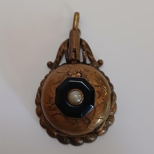 May include: A gold locket pendant with an ornate design. The locket has a black cameo with a white pearl in the center.