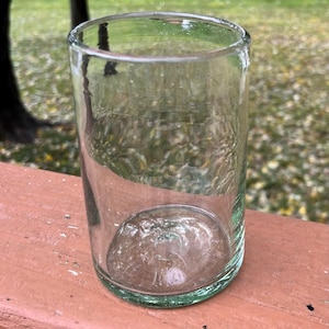 May include: A clear, cylindrical drinking glass with a rounded bottom. The glass is made of transparent material, allowing visibility of the contents. The glass sits on a brown surface, with a blurred green background.