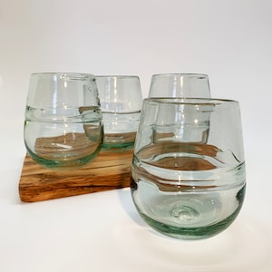 May include: Four clear, stemless drinking glasses with a rounded shape. The glasses are arranged on a wooden cutting board. The glasses have a slight green tint and a textured, handcrafted appearance. The background is white.