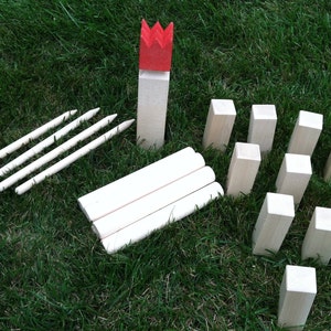 May include: A set of wooden lawn bowling game equipment. The set includes 10 white wooden pins, 3 white wooden throwing sticks, and a red crown-shaped wooden top piece.