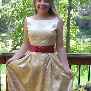 May include: A woman wearing a long, cream-colored satin dress with a red sash and floral pattern. She is standing on a wooden deck with a railing and a potted plant in the background.