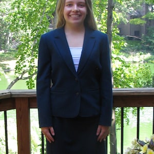 May include: A woman wearing a dark blue blazer and skirt suit with a white blouse. She is standing on a wooden deck with a railing and green foliage in the background.