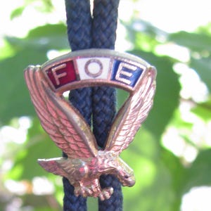 May include: A vintage bolo tie featuring a bronze eagle with outstretched wings. The eagle is perched above a banner with the letters "FOE" in red, white, and blue. The tie has a navy blue cord with silver-toned tips.