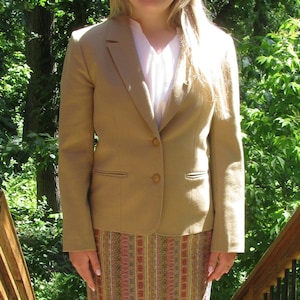 May include: A woman wearing a tan blazer with a white shirt underneath and a multi-colored striped skirt.