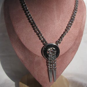 May include: A silver-toned necklace with a large circular pendant and two dangling rectangular bars. The necklace is displayed on a pink velvet bust form, showcasing its design and style. The chain is made of thick, rounded links.